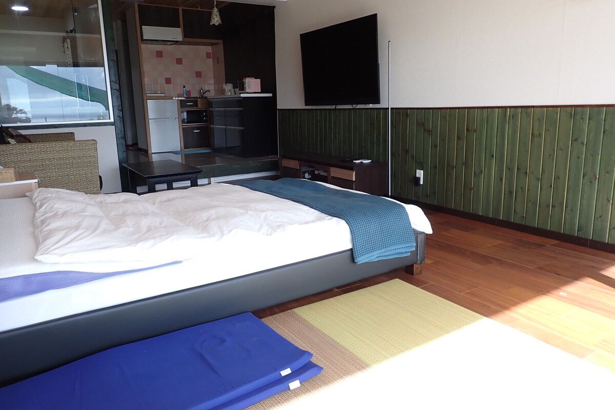 A bedroom with a bed, a small desk, a flat-screen TV, and a kitchenette area.