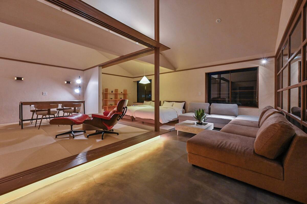 A modern living room with a sofa, a red chair, a dining table with chairs, and a large window.
