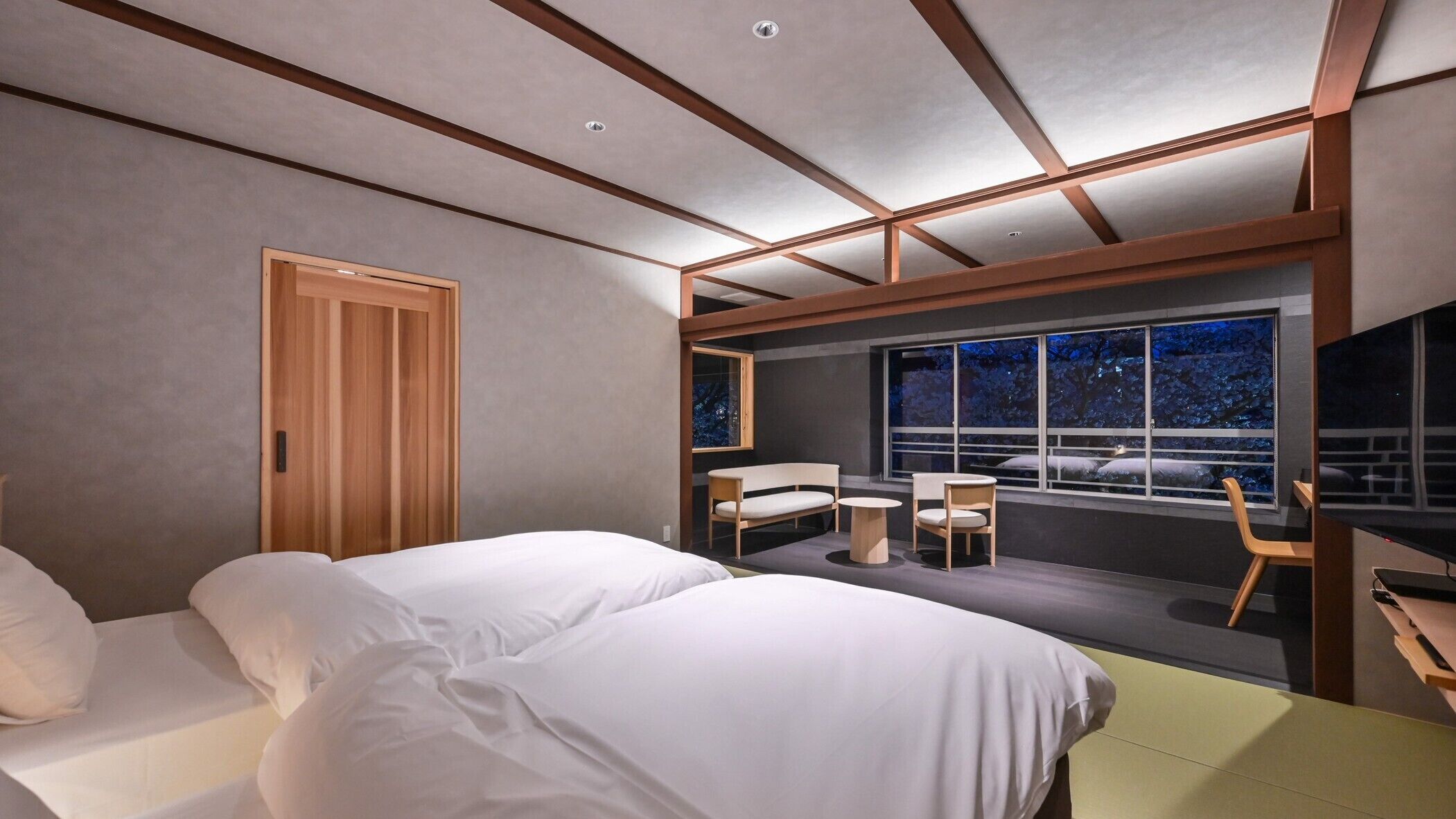 Une chambre d’hôtel moderne équipée d’un lit, d’un bureau et d’une grande fenêtre offrant une vue sur la ville la nuit.
