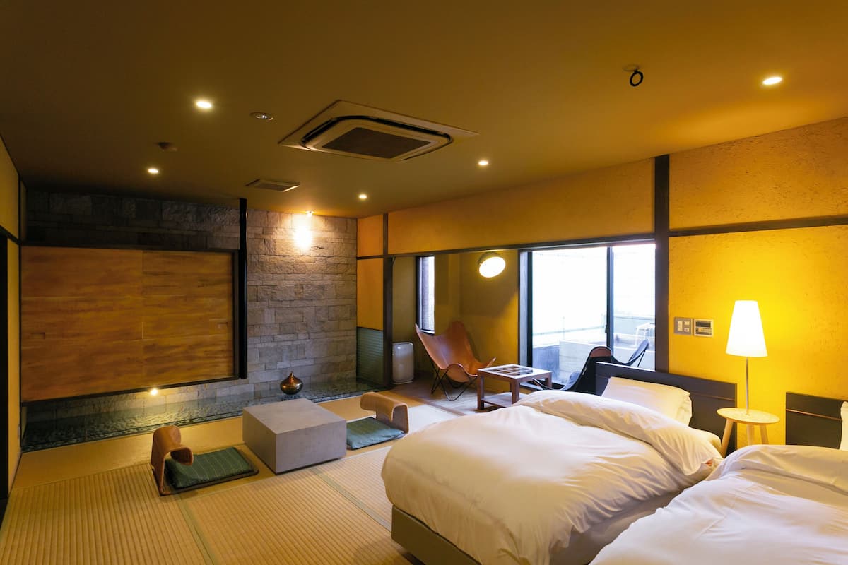 A modern hotel room with a large bed, a seating area with a chair and a small table, a stone accent wall, and large windows.