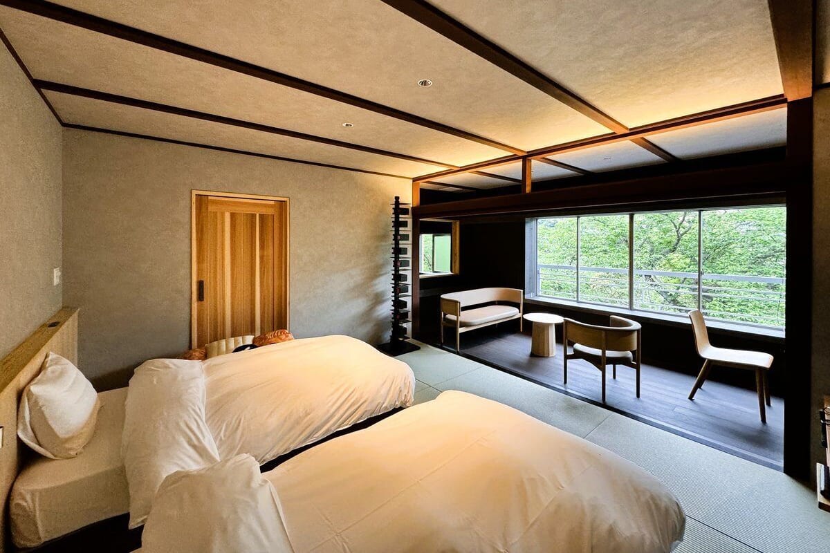 A modern hotel room with two beds, a wooden wardrobe, a small table, and a large window offering a view of greenery.