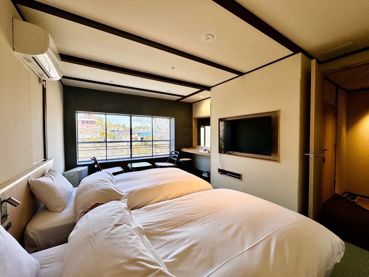 A hotel room with a large bed, a TV mounted on the wall, and a window with a view of buildings outside.