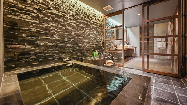 Un área de spa con una pared de piedra, una bañera de hidromasaje y una puerta corredera de madera que da a un baño.