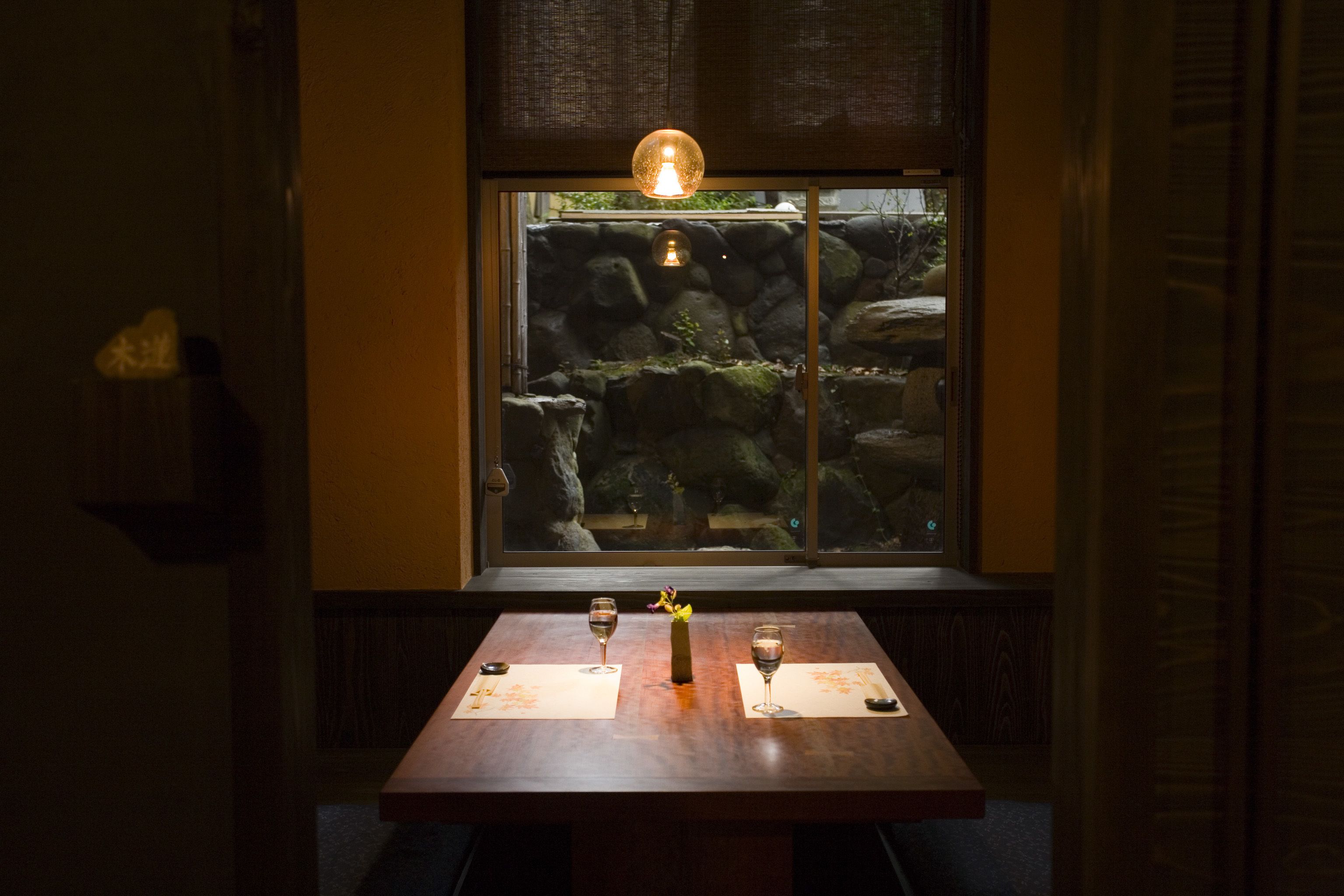 A table with two wine glasses and a vase, set against a window with a view of rocks and greenery.
