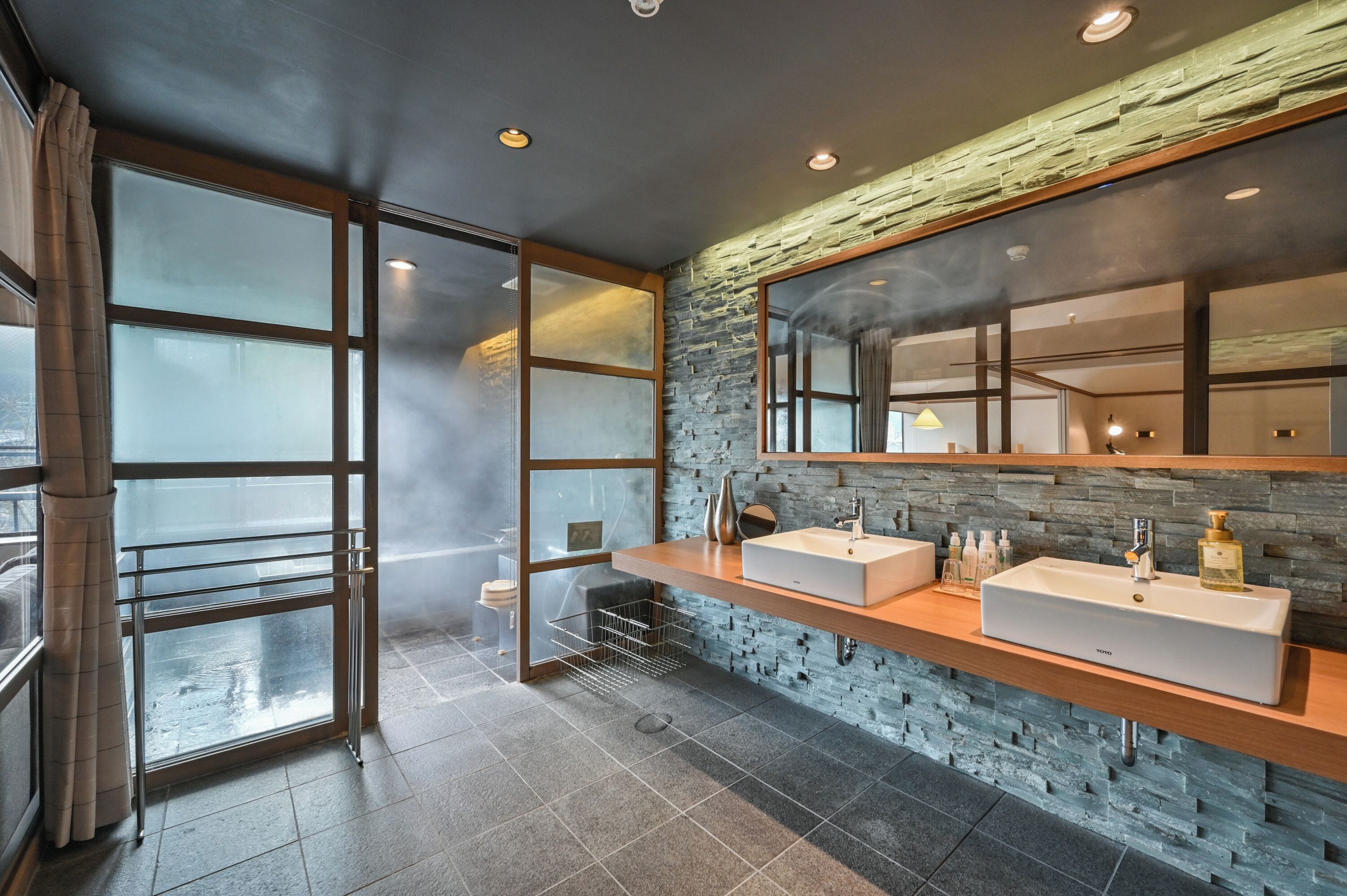 A modern bathroom with two sinks, a large mirror, and a stone wall.