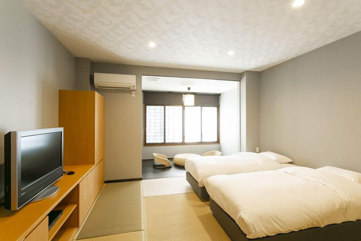 A hotel room with a TV on a wooden stand, a bed with white bedding, a window with blinds, and an air conditioning unit on the wall.