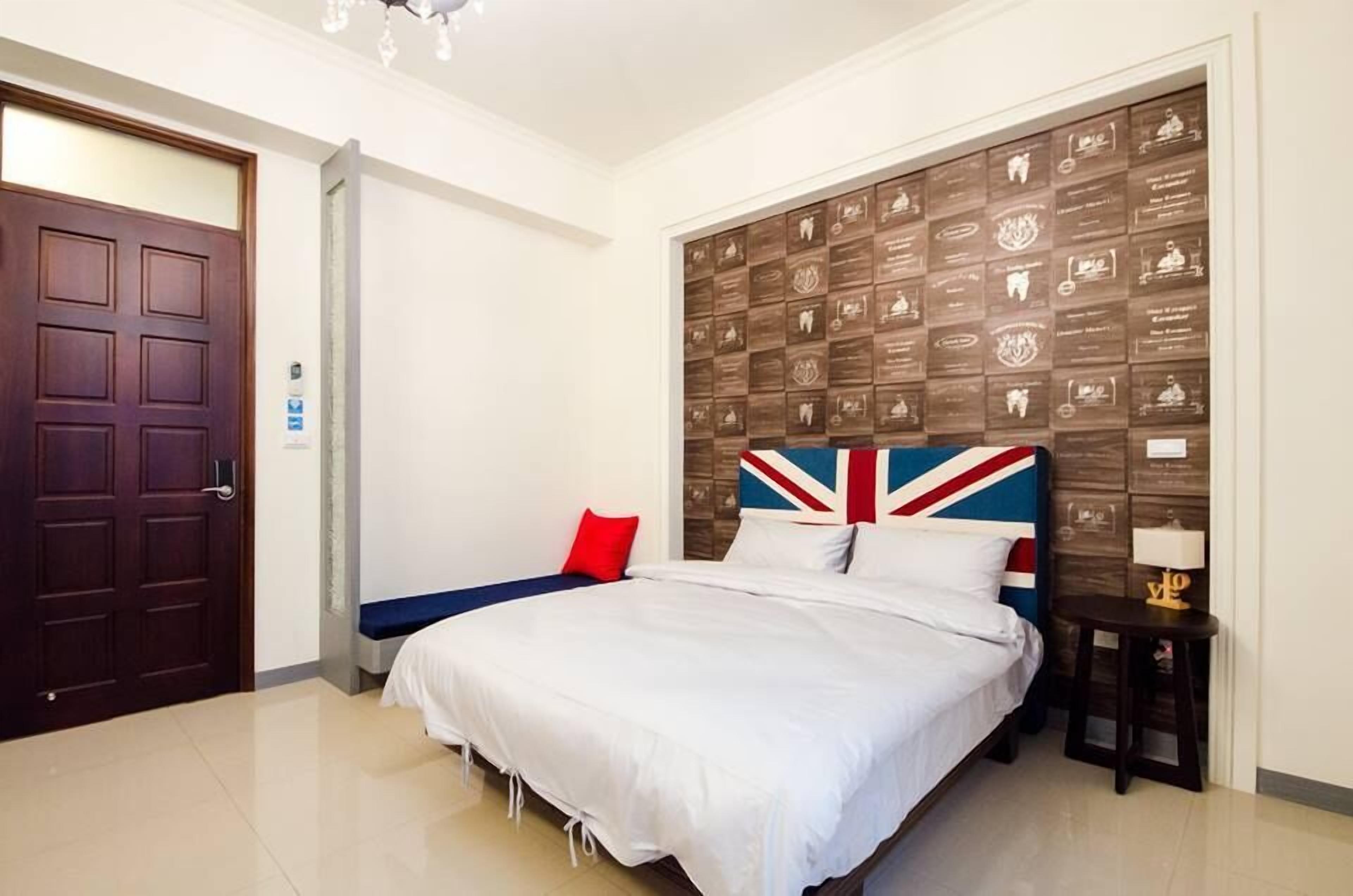 A hotel room with a large bed, a wall-mounted plaque display, a red pillow, and a small bedside table with a lamp.