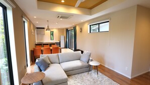 A modern living room with a sectional sofa, a small round table, and a kitchen in the background. - Tabinoteitaku Okinawa Bise Homann Concept (Motobu)