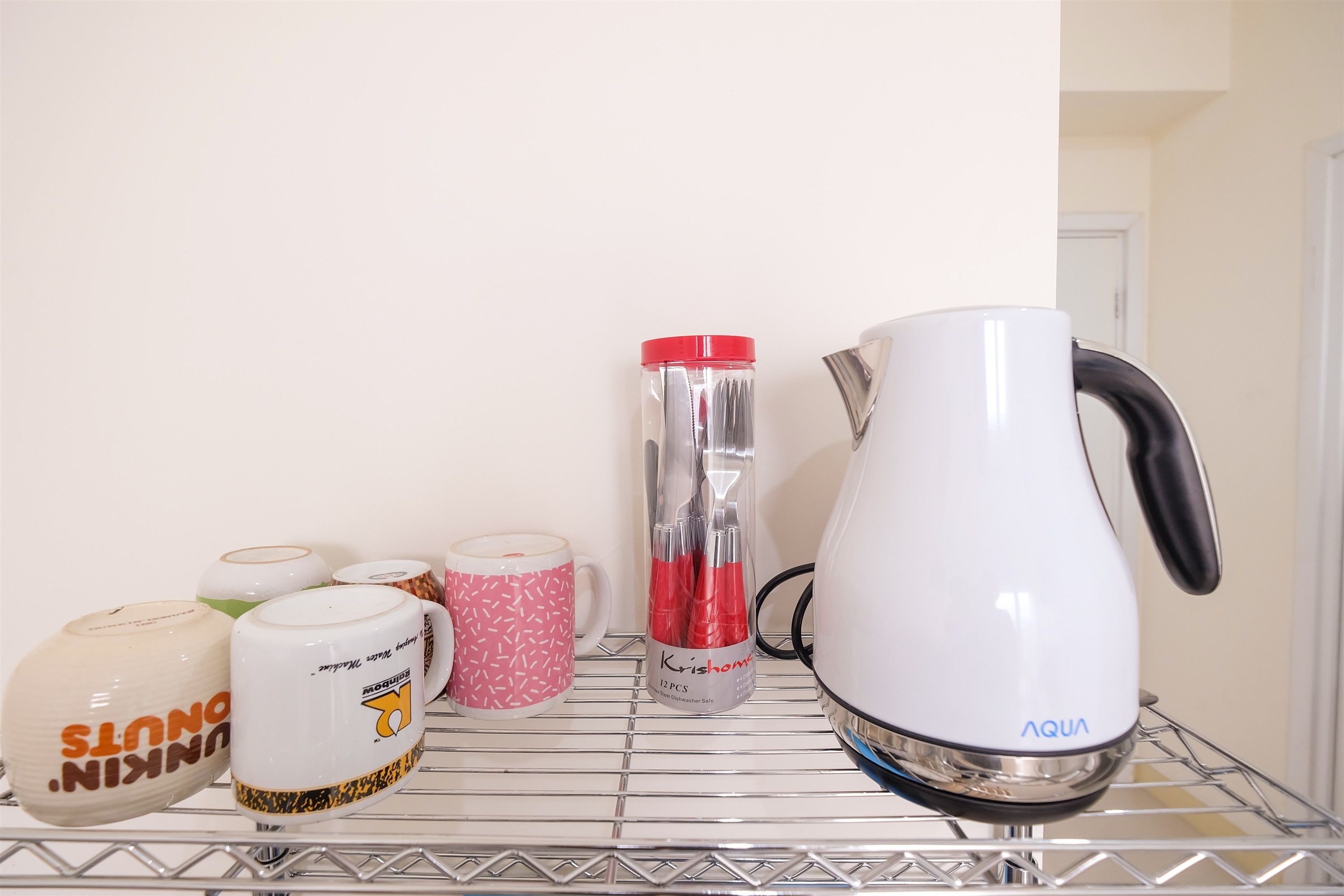 A kitchen shelf with various kitchenware items including a white kettle, a pink mug, and a clear container with utensils.