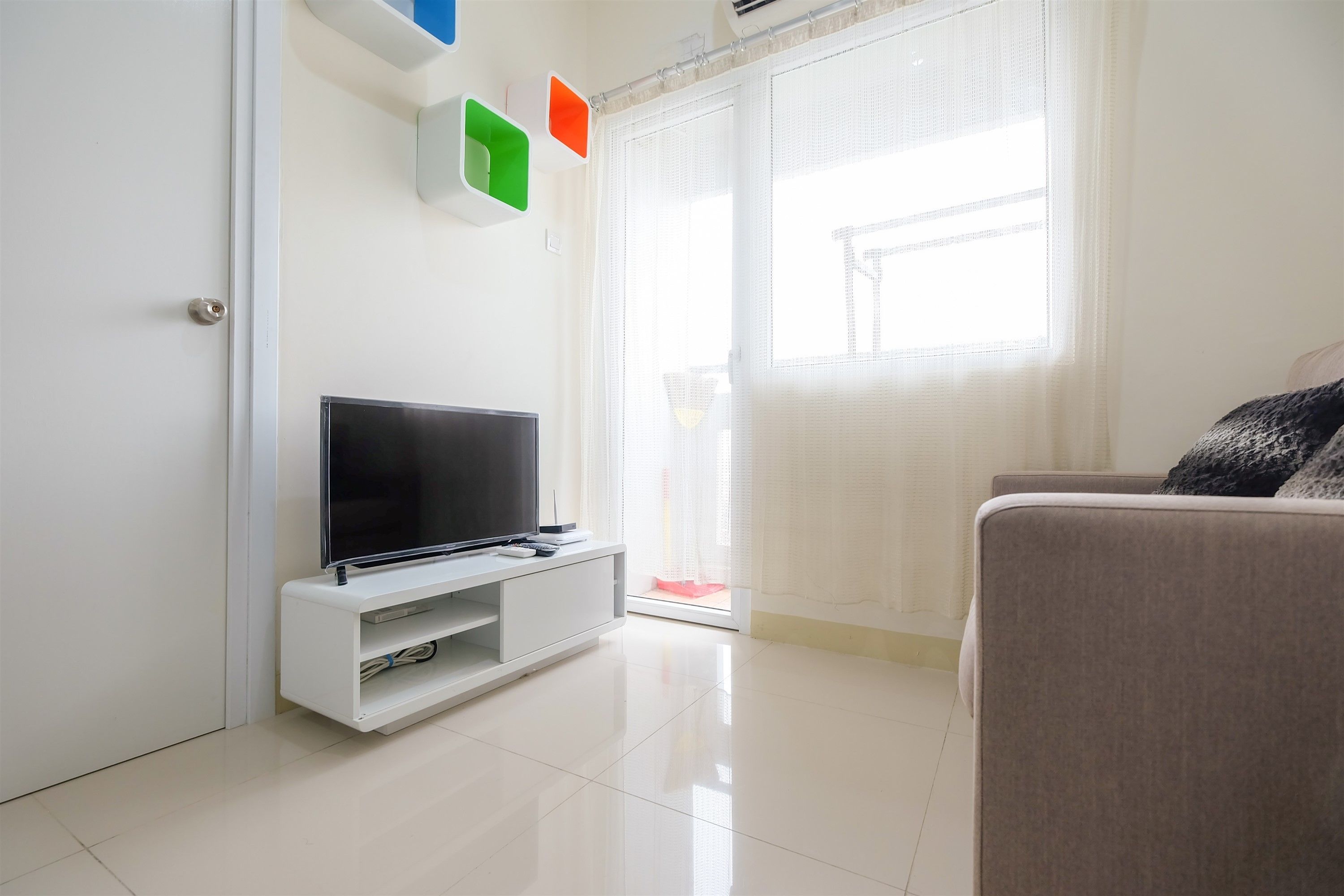 Una sala de estar moderna con un televisor de pantalla plana sobre un soporte blanco, un sofá color beige y un ventanal corredizo que da a un balcón.