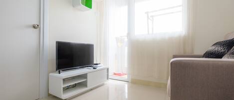 Ein modernes Wohnzimmer mit einem Flachbildfernseher auf einem weißen Schrank, einem beigefarbenen Sofa und einer Glastür, die auf einen Balkon führt.
