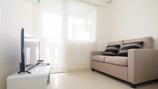 Un salón con un televisor de pantalla plana sobre un soporte blanco, un sofá beige con dos cojines estampados y un ventanal corredizo que da a un balcón.
