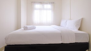 Una cama bien hecha con sábanas blancas y una toalla enrollada, una ventana con cortinas translúcidas y un cabecero negro.
