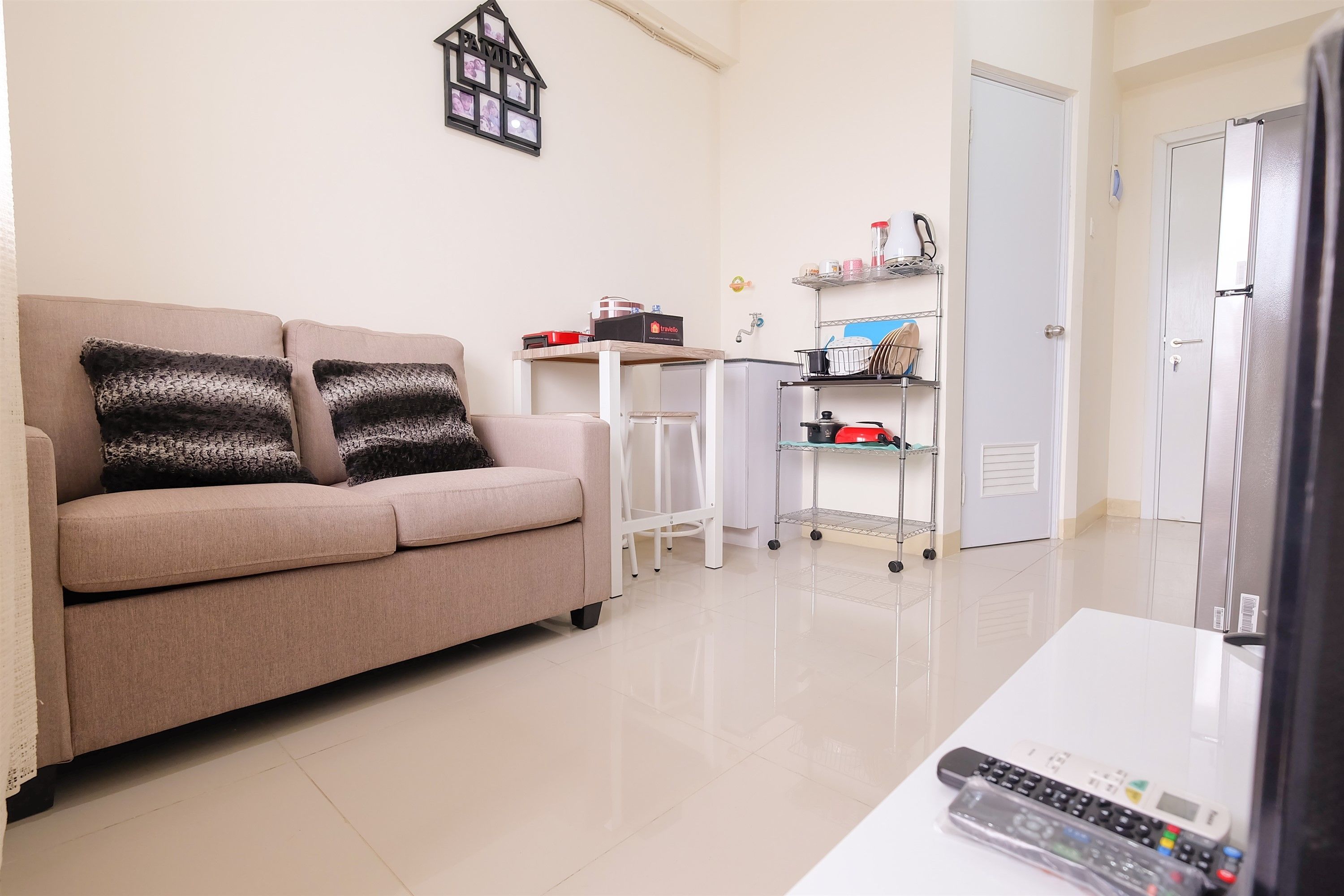A living room with a beige sofa, two black patterned pillows, a white table with a remote control, a small white table, and a kitchen area with shelves and appliances.