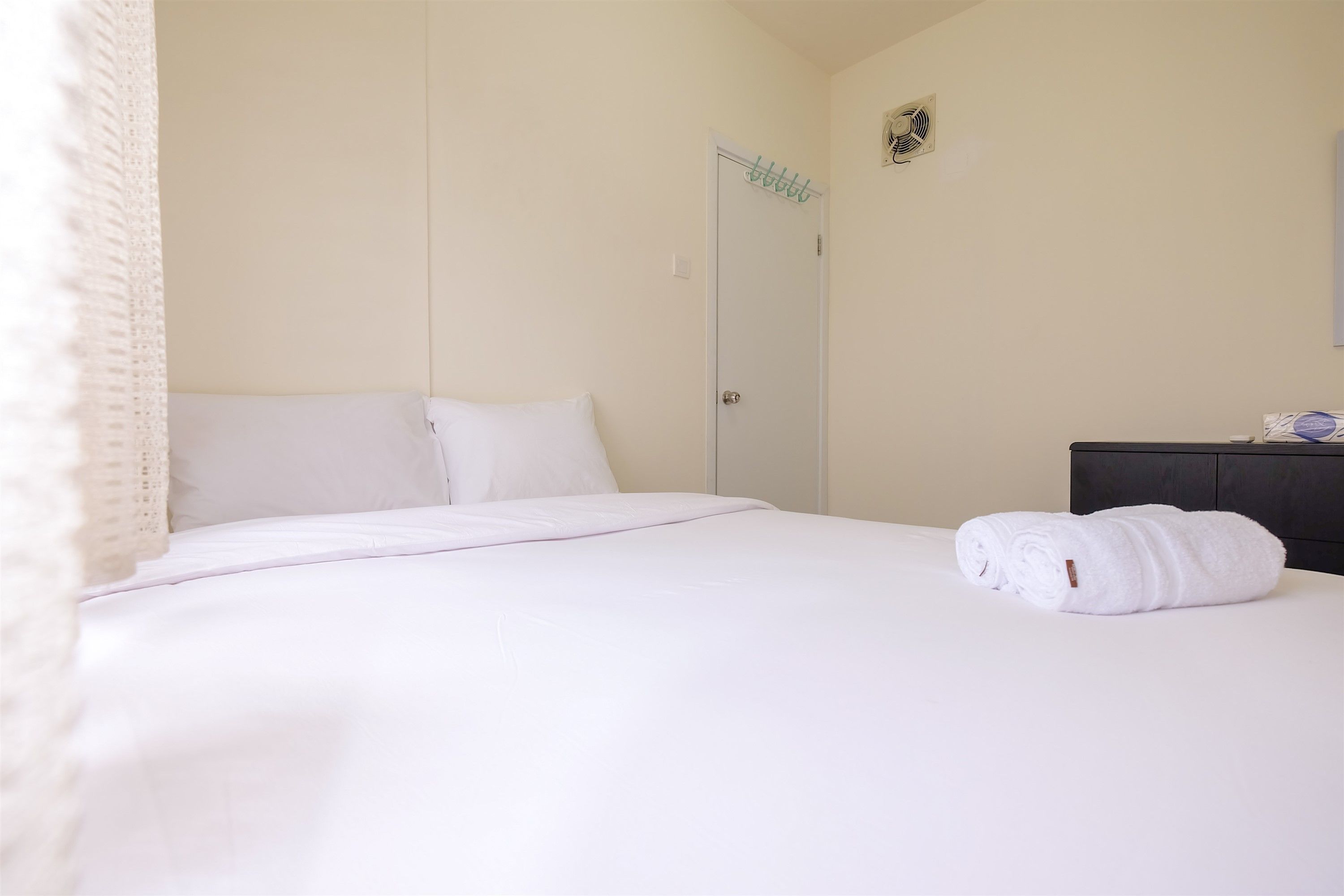 Ein ordentlich bezogenes Bett mit weißen Leinen, eine dunkle Kommode und eine geschlossene Tür mit einem kleinen Ventilator an der Wand.