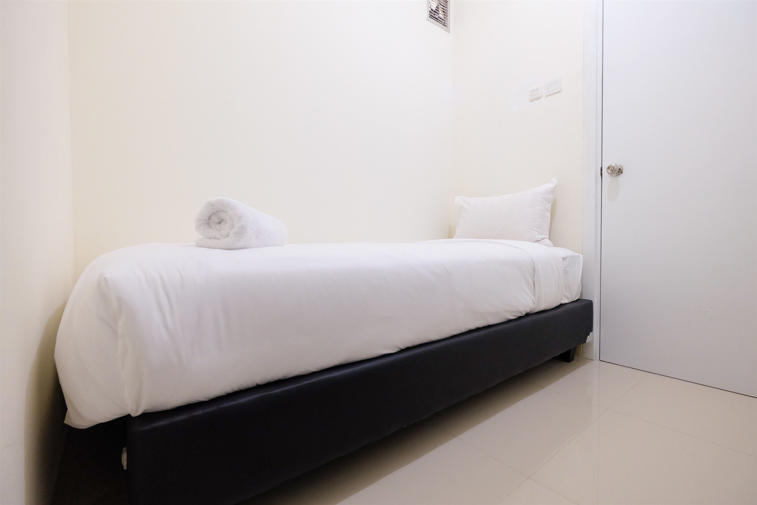 A single bed with white bedding and a rolled towel, a black headboard, and a white door in a minimalistic room.