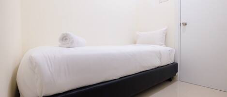 A single bed with white bedding and a rolled towel, a black headboard, and a white door in a minimalistic room.