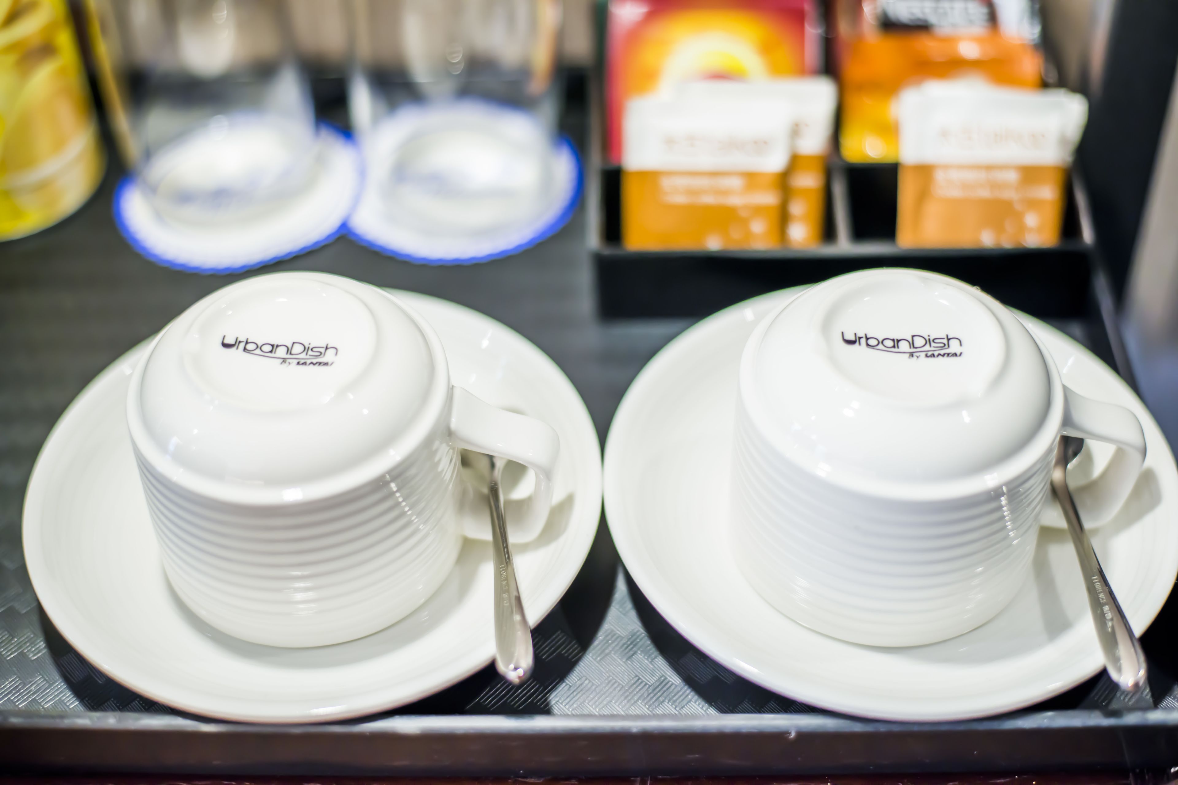 Two white ceramic cups with lids and saucers, each with a spoon, labeled "UrbanDish by Lanna."