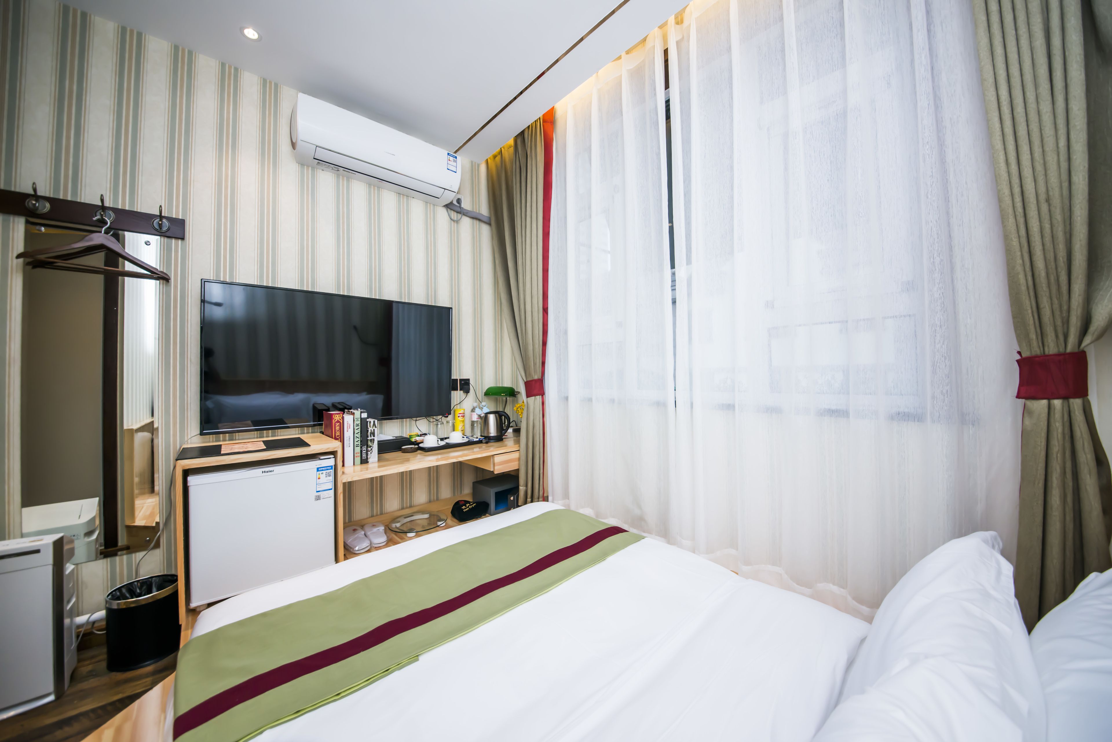 A hotel room with a bed, a desk with a television, a small refrigerator, and a window with curtains.