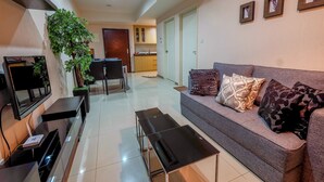 A modern living room with a grey sofa, black coffee table, and a TV on a black stand. There is a large green plant and a wooden cabinet in the background. - 1 Bedroom Apartment Casa Grande Residence by Travelio (Jakarta)