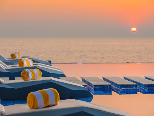 Fileira de espreguiçadeiras com toalhas listradas, vista clara do oceano e um pôr do sol ao fundo.