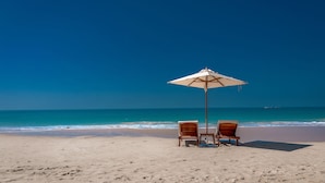 Una playa con dos sillas de madera y un paraguas grande bajo un cielo azul despejado.