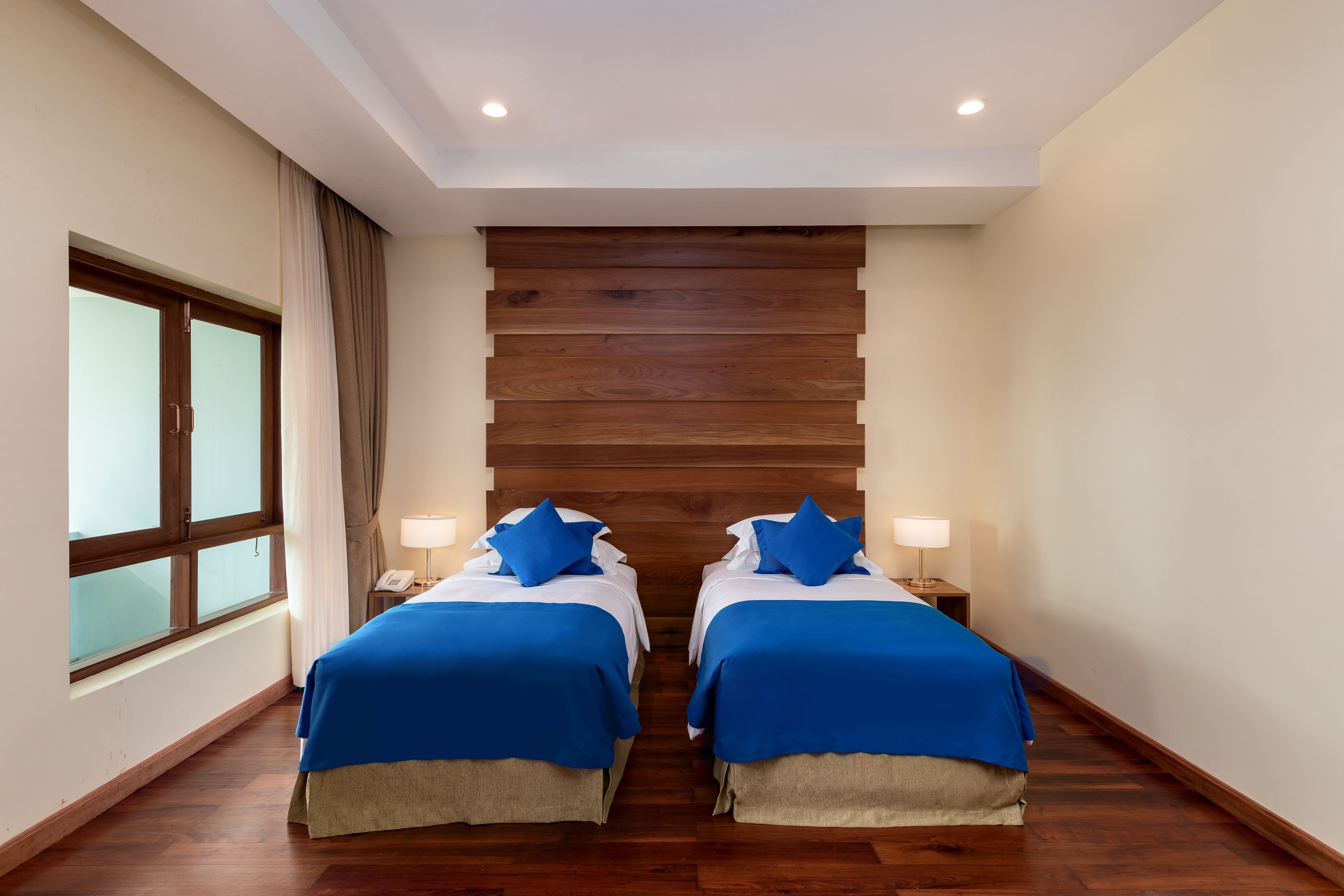 Habitación de hotel con dos camas, cabecero de madera, lámparas de noche y un ventanal con cortinas.