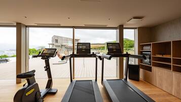 A modern gym with three treadmills, a stationary bike, and a weight bench. Large windows offer a view of a cityscape and greenery.