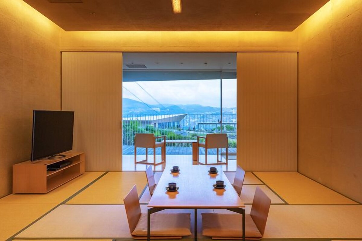 A room with tatami mats, a table with chairs, a television, and a view of a bridge and mountains.