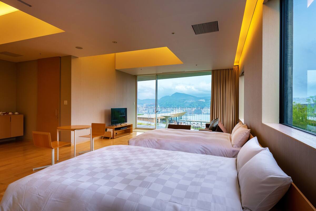 A hotel room with two beds, a TV, a desk, and a large window with a city view.