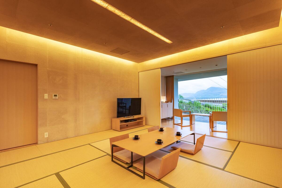 A modern hotel room with tatami flooring, a flat-screen TV, a dining table, and a balcony with a view of the mountains.