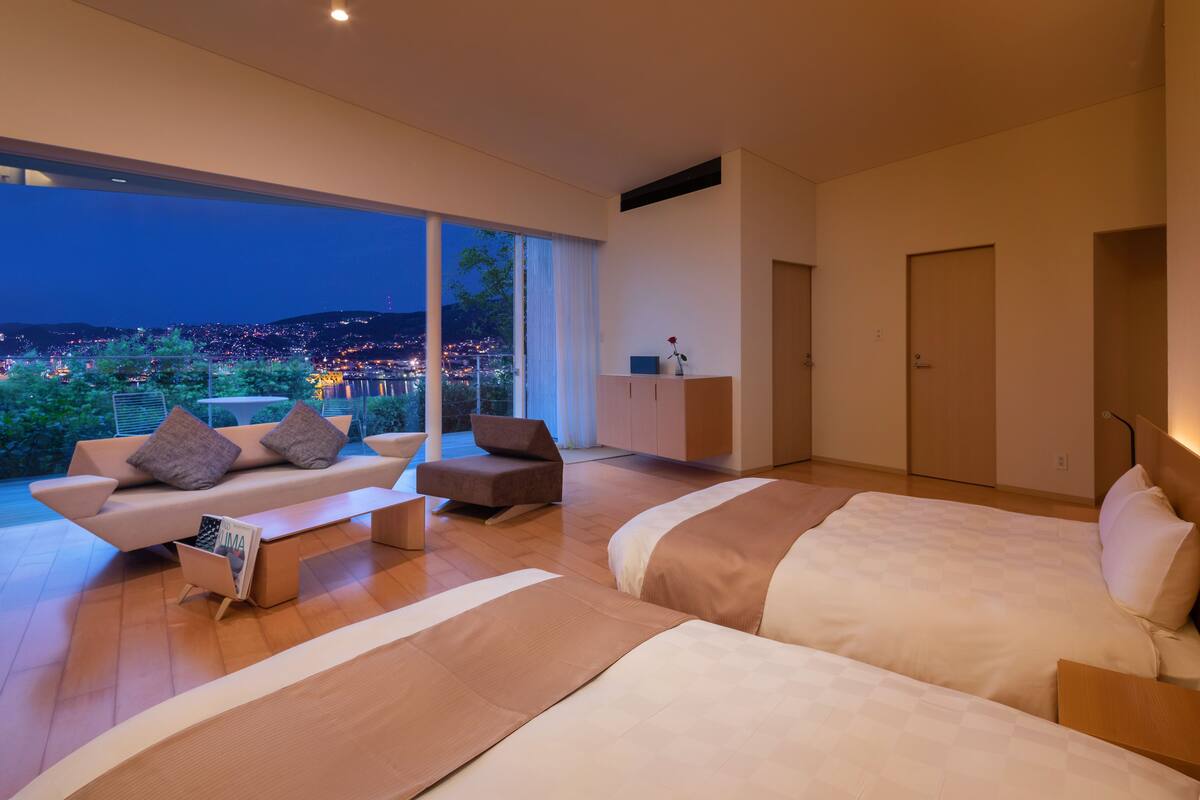 A modern bedroom with a large bed, a sofa, and a chair. There is a view of a cityscape through a large window.