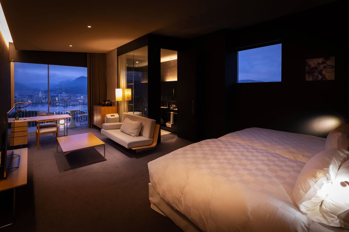 A modern hotel room with a large bed, a sofa, a desk, and a view of a cityscape through large windows.