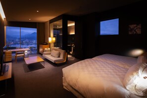 A modern hotel room with a large bed, a sofa, a desk, and a view of a cityscape through large windows. - Garden Terrace Nagasaki Hotels & Resorts (Nagasaki)