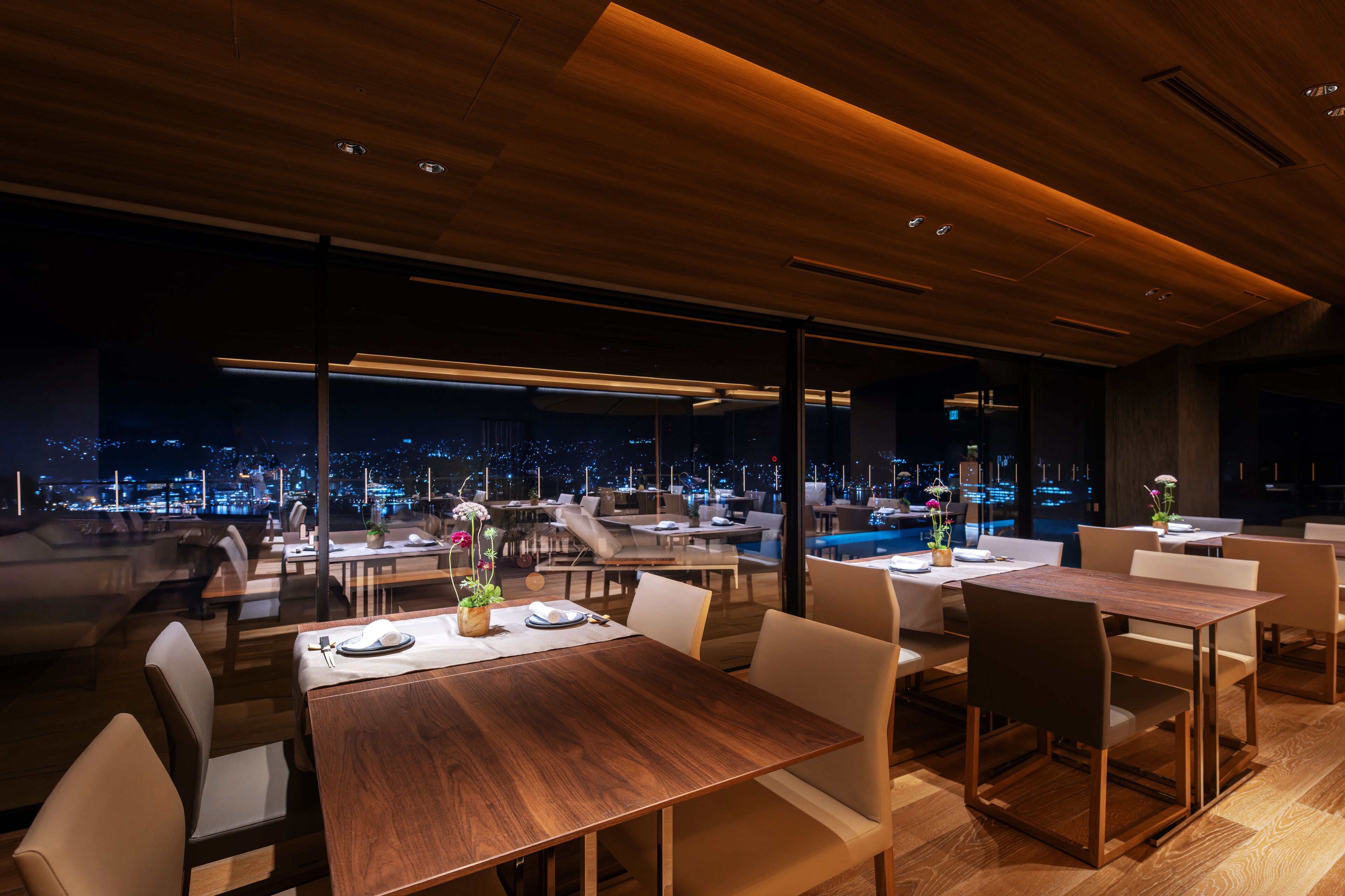 Een modern restaurant met houten tafels en stoelen, grote ramen met uitzicht op de stad 's avonds.