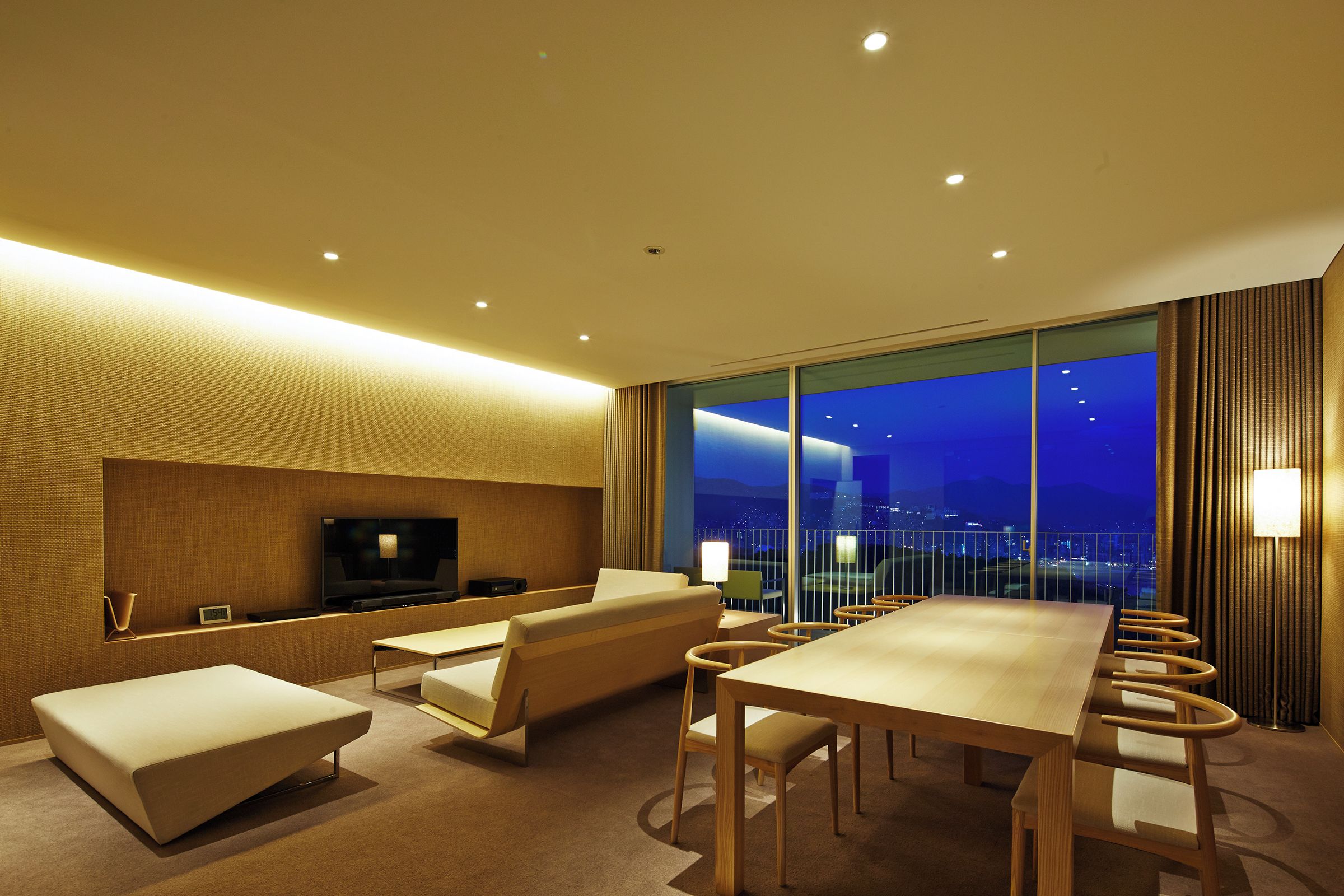 A hotel room with a large window, a flat-screen TV, a sofa, a dining table, and a view of the city at night.