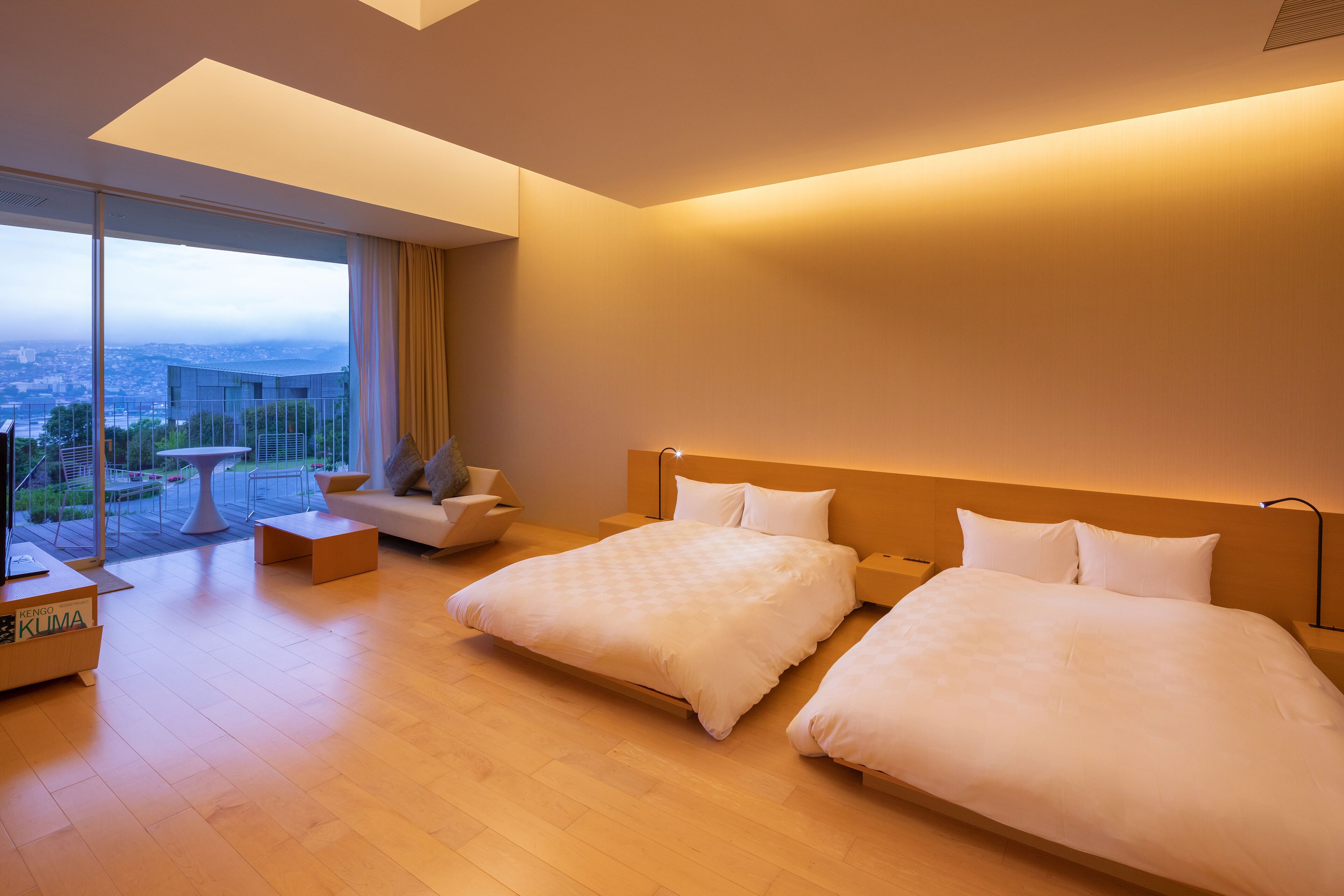 A modern hotel room with two beds, a sofa, a small table, and a large window offering a city view.