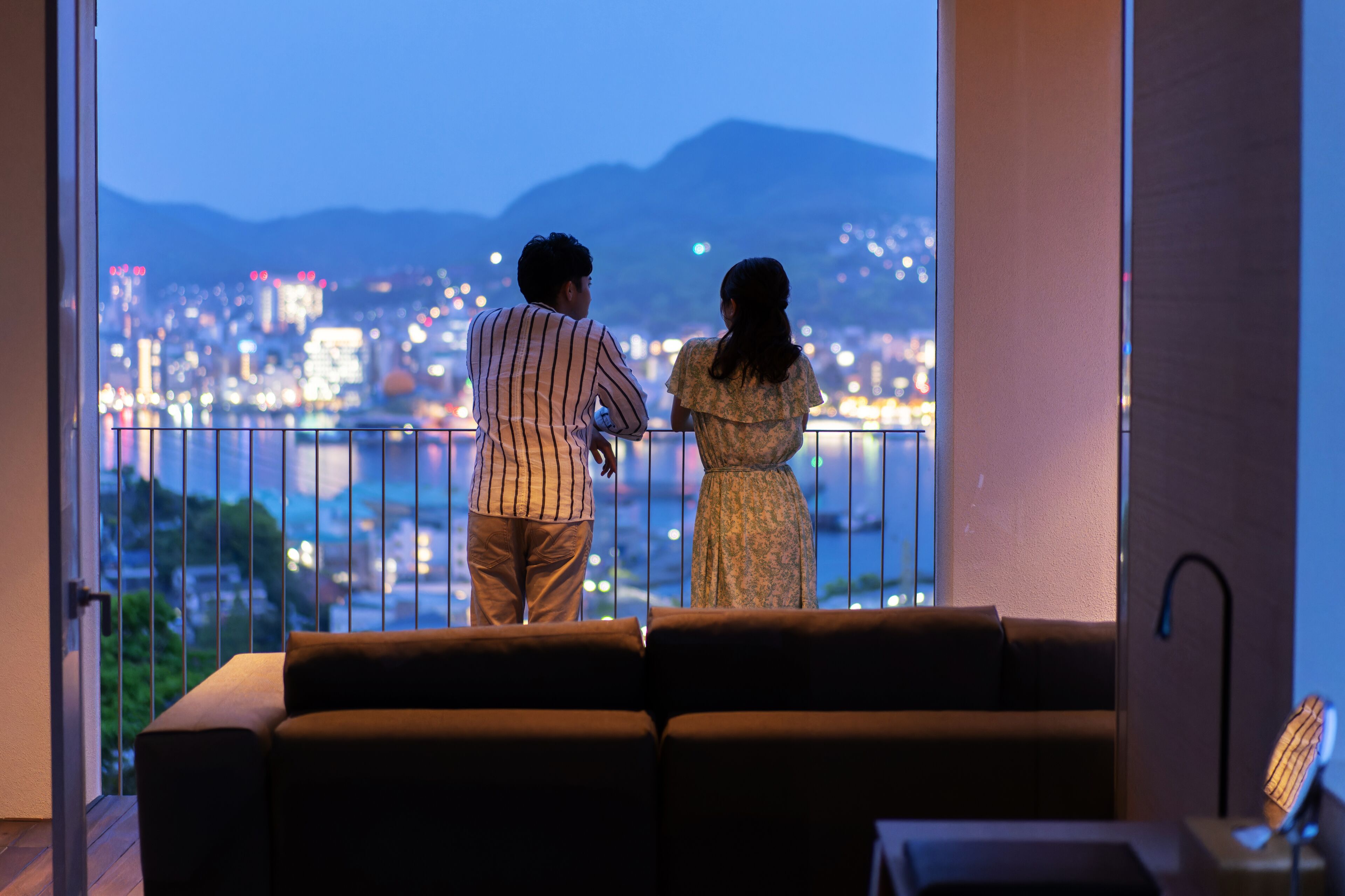 A couple standing on a balcony overlooking a cityscape with mountains in the background.