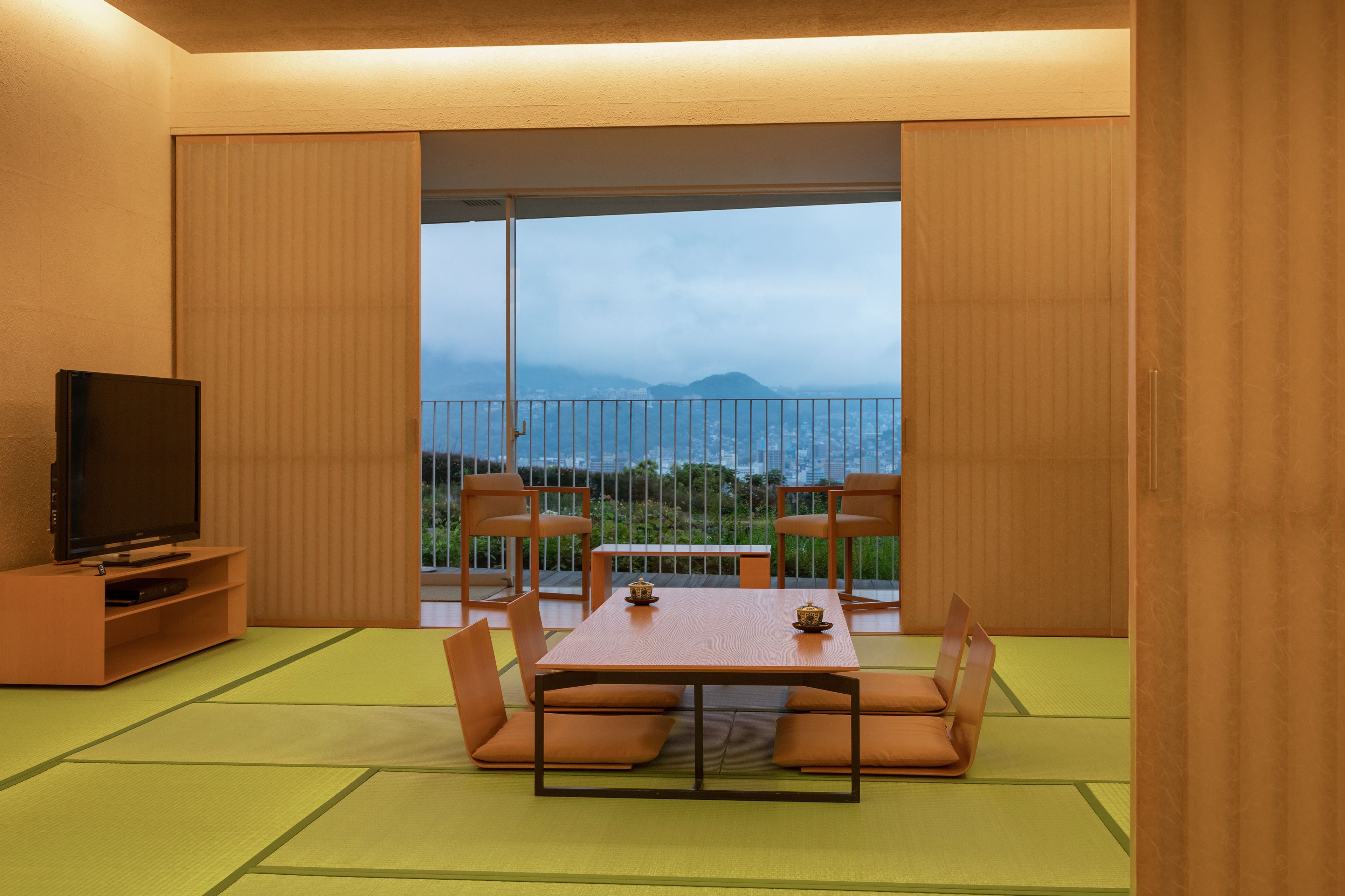 Quarto com piso de esteiras de tatame, mesa baixa com almofadas, televisão e varanda com vista para montanhas.