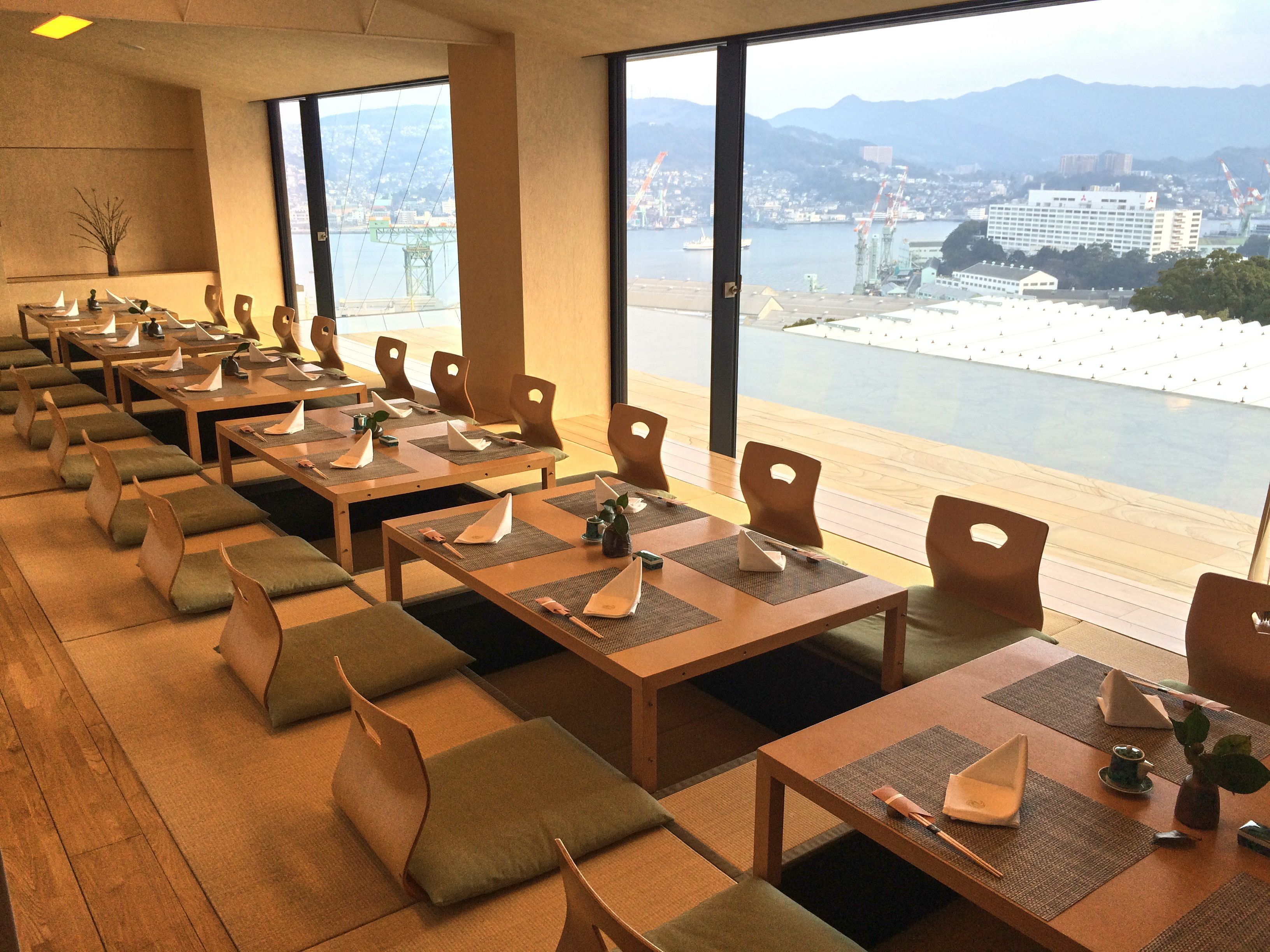 A restaurant with long tables and chairs, large windows with a view of a waterfront and mountains.