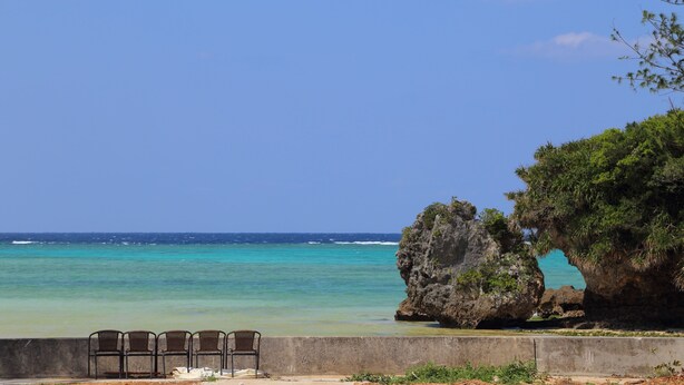 A coastal view with a concrete barrier, three metal chairs, and a large rock formation with a natural archway. - Tabinoteitaku OkinawaNakijin HOMANN CONCEPT (Nakijin)
