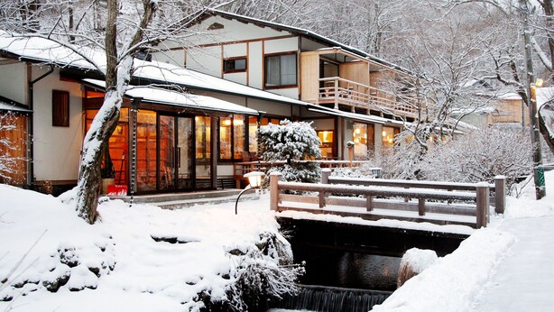Un bâtiment de style japonais traditionnel, avec un pont en bois enjambant un ruisseau, le tout entouré d’arbres couverts de neige.