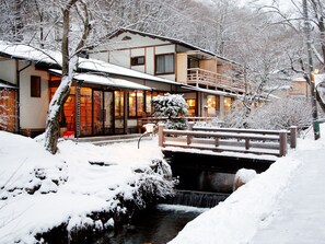 A traditional Japanese-style building with a wooden bridge over a stream, surrounded by snow-covered trees.