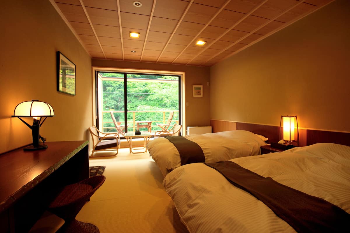 A hotel room with two beds, a desk with a lamp, a chair, and a view of greenery through the window.