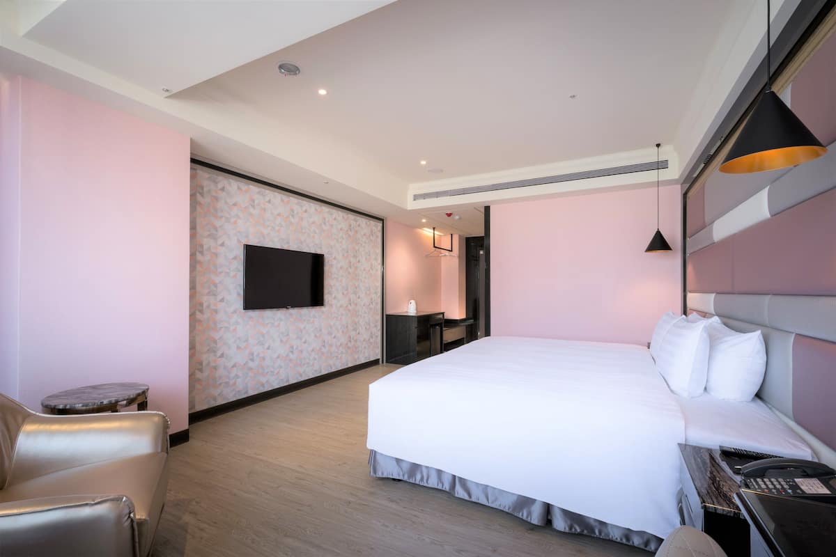 Ein modernes Hotelzimmer mit einem großen Bett, einem Schreibtisch mit Stuhl, einem flachbildfernseher an der Wand und einer kleinen Sitzecke mit Sessel und Tisch.