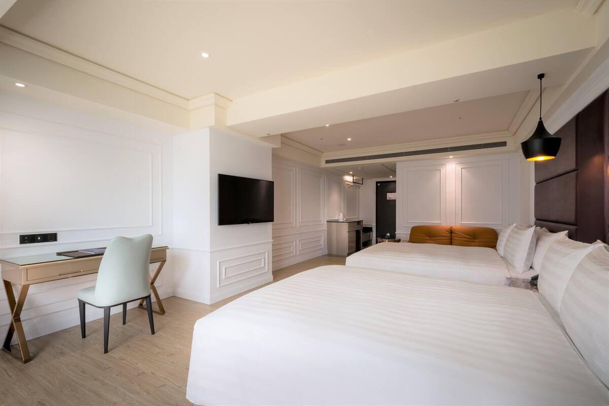 Ein Hotelzimmer mit zwei Betten, einem Schreibtisch, einem Sessel, einem Fernseher und einer modernen Lampe.