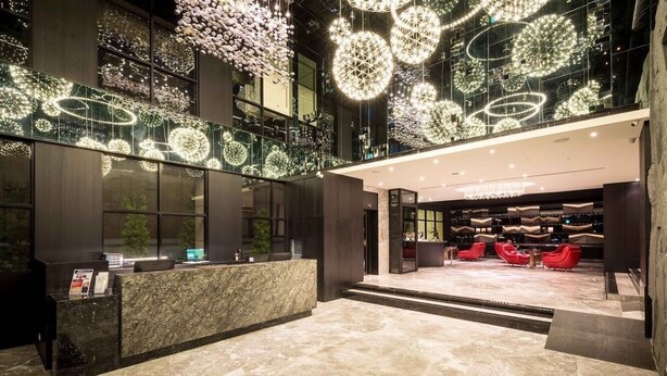 Un hall d’entrée moderne avec un comptoir d’accueil en marbre, un espace détente avec des sièges rouges et des luminaires décoratifs au plafond.
