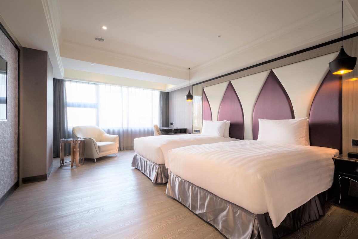 Ein Hotelzimmer mit zwei Betten, einer Sitzecke mit einem Sessel, einem kleinen Tisch und einem großen Fenster, das für natürliches Licht sorgt.
