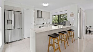 Eine moderne Küche mit Edelstahl-Kühlschrank, weißen Schränken, einer großen Kochinsel mit Sitzgelegenheiten und Blick durch ein Fenster.