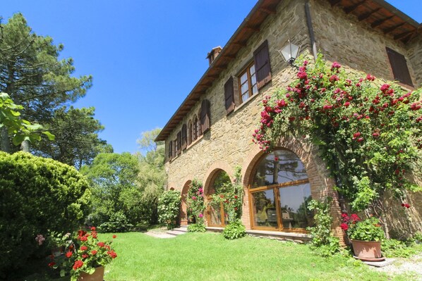 A stone building with arched windows and a garden with flowering plants.